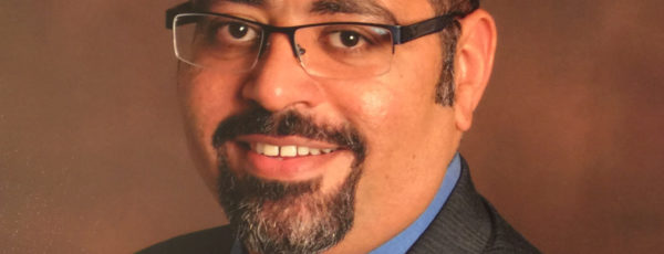 Dr. Ehab Hanna standing in front of a brown background. He has short, dark hair and is wearing a black suit and glasses.