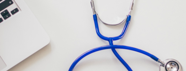 An open laptop with black keys sits next to a bright blue stethoscope on a white surface.