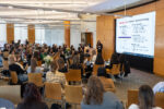 A large group of people attending a conference or seminar in a modern conference room. A presenter stands near a screen displaying information on "Learning Curve: Technical + Business Strategy."