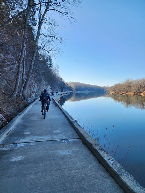 A group of cyclists ride along a paved path beside a calm river on a clear, sunny day. The path curves gently along the water, with bare trees and rocky cliffs lining one side and the serene river reflecting the blue sky and surrounding landscape.