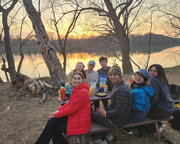 A group of seven people sit around a picnic table enjoying snacks and drinks at sunset by a calm river. They are dressed in jackets and warm clothing, smiling at the camera, with bare trees and a scenic, golden sky reflected in the water behind them.
