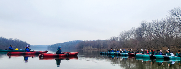 A group of people in colorful kayaks float on a calm river surrounded by leafless trees on an overcast day. The paddlers are wearing jackets and hats, suggesting cool weather, and are split into two groups facing each other near the riverbank.