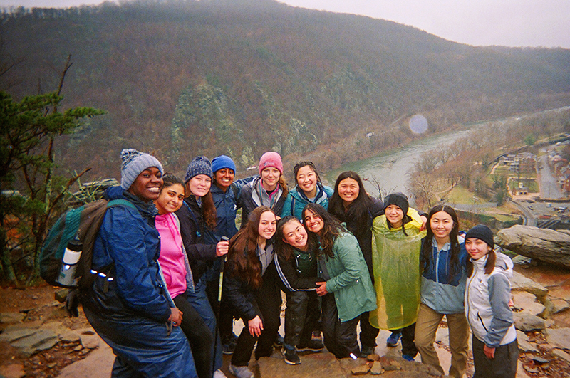A group of smiling hikers pose together on a rocky overlook with a river winding through a valley below. The hikers are dressed in outdoor gear and rain jackets, suggesting wet weather, and the background features misty hills covered in bare trees, indicating early spring.