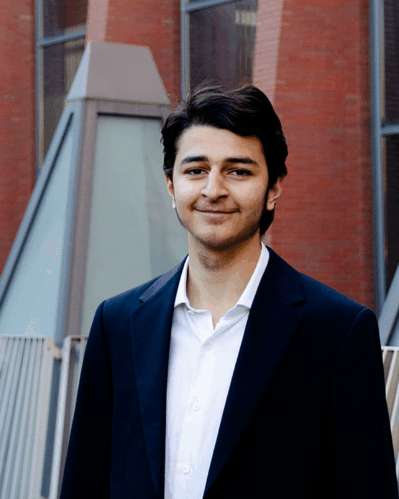 Young man in a white dress shirt and dark blazer smiling in front of a red brick building.