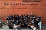 A large group of people in matching Wharton School t-shirts posing in front of a building sign that reads "The Wharton School, University of Pennsylvania, Jon M Huntsman Hall."