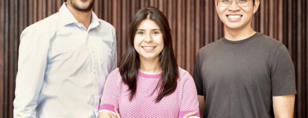 A group of three people smiling, standing close together in front of a vertical wooden slat background. They appear to be in a casual setting.