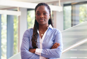 Headshot of person with crossed arms, wearing a light blue shirt and gold jewelry, standing indoors with blurred background.