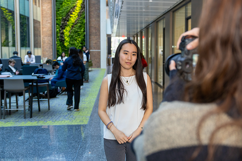 A young woman stands posing for a photo in a modern indoor atrium, while a photographer captures her image. Behind them, students sit and walk through the bright space featuring tall glass windows and a vertical green plant wall
