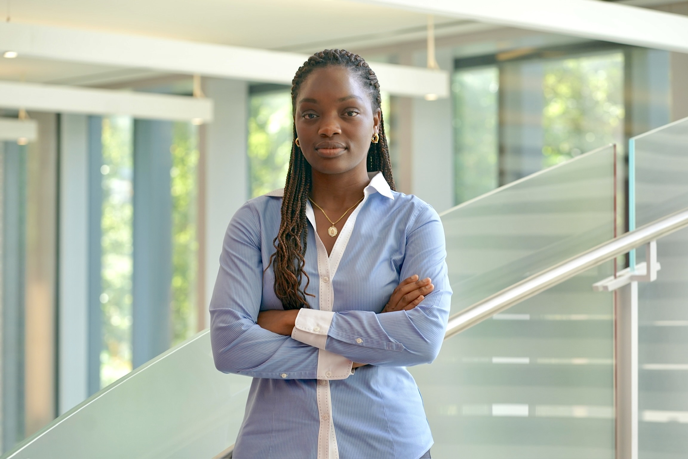 Headshot of a person standing with arms crossed in a professional setting, wearing a blue shirt and gold jewelry.