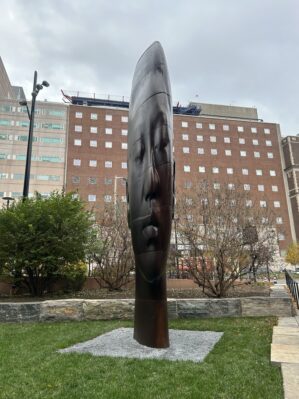 An oversized sculpted bust of a woman's head stands outside the Penn Museum with hospital buildings in background.