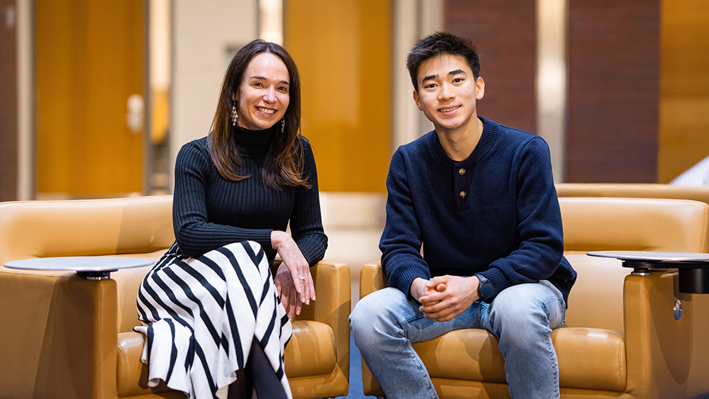 Two people sitting in tan chairs, smiling toward the camera in a bright indoor setting.