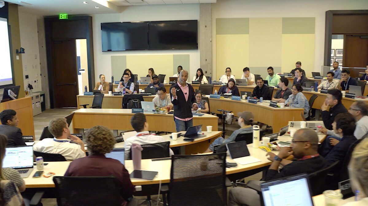 Americus Reed addresses a classroom of students, standing in the center of a semicircle seat arrangement.