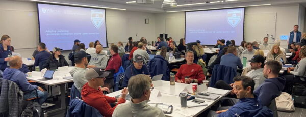 Penn athletics coaches sit and confer at tables in a busy classroom.