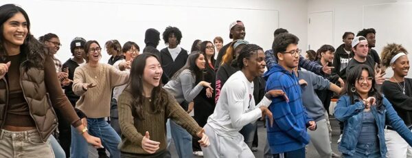 A large group of people, diverse in appearance, are standing in a room participating in a dance class or activity, smiling and moving together.