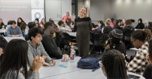 Two professors (center and background), stand amidst a classroom of students seated at tables.